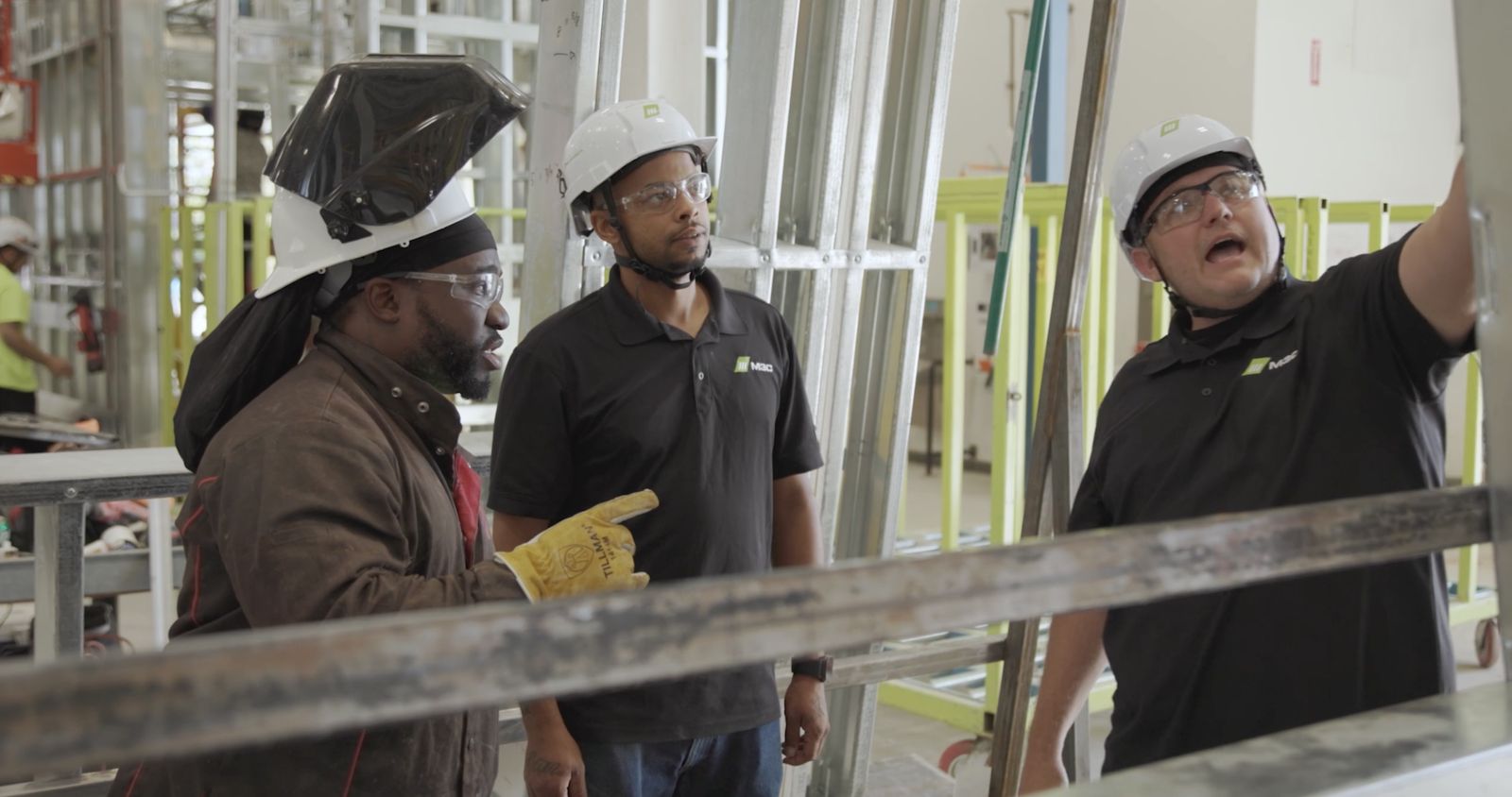 Three Mark III team members in hard hats and safety glasses talk through a steel frame on the factory floor.