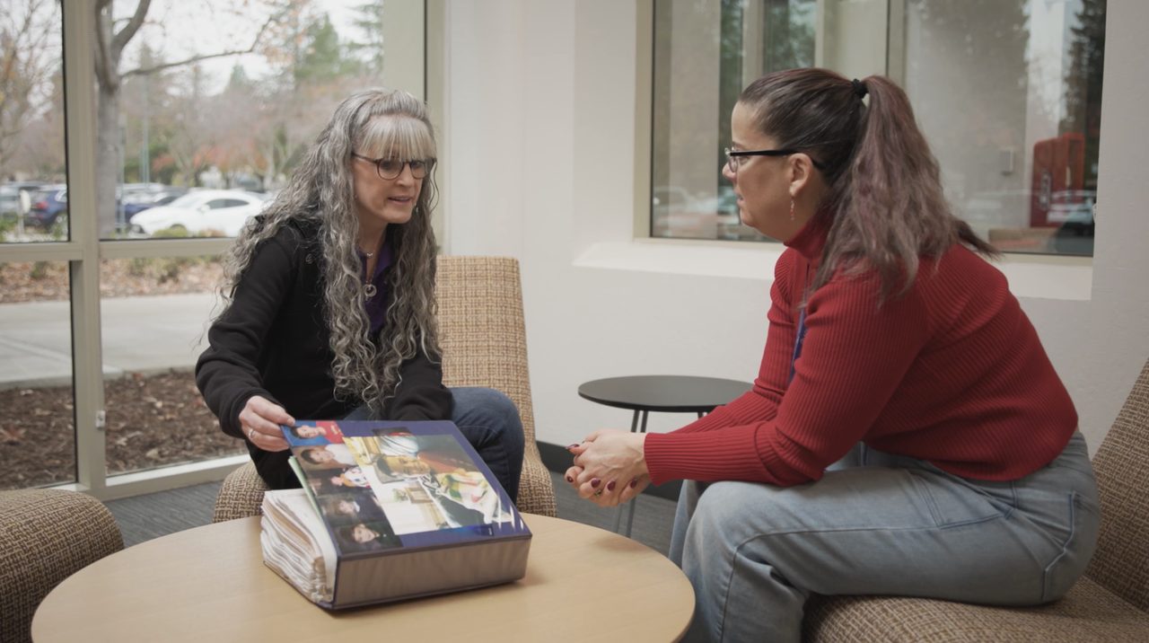 Two women talk across a small table in a sunlit lobby, one turning the pages of a thick binder of family photos.