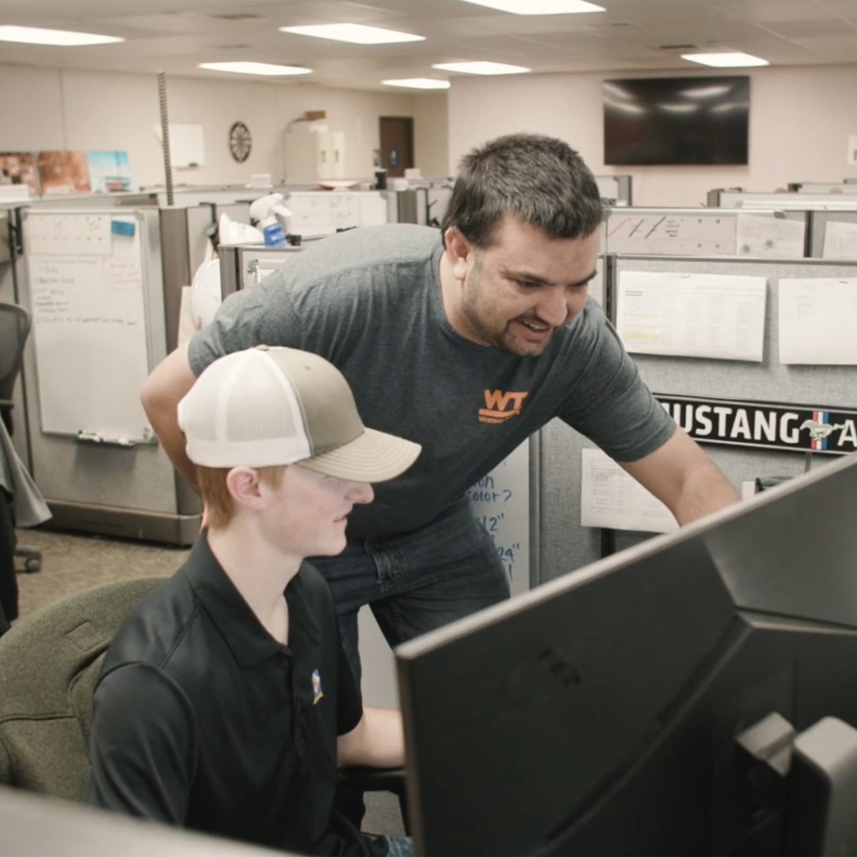 A young new hire at his desk as a senior teammate leans in to look at his screen with him.